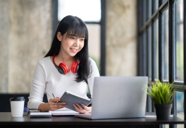 Happy Asian girl student wear wireless headphone study online with the teacher, happy young woman learn language listen lecture watch webinar write notes look at laptop sit in cafe, distance education