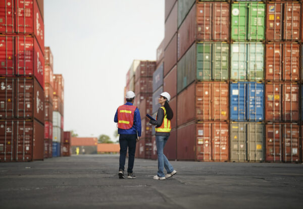 Two workers in safety gear review plans and discuss operations in a container yard.